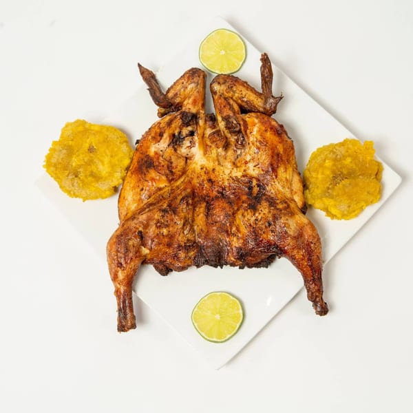 Whole grilled chicken, split and flattened, served with two tostones and two lime slices on a white plate.