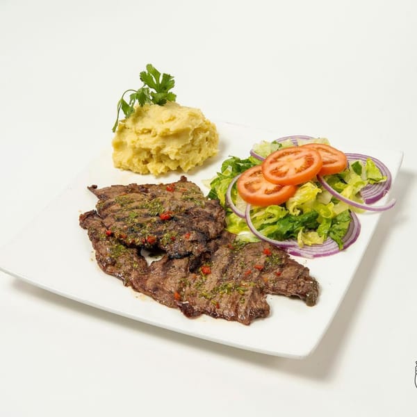 Grilled steak with chimichurri sauce, mashed potatoes, and a side salad with tomatoes and red onions.